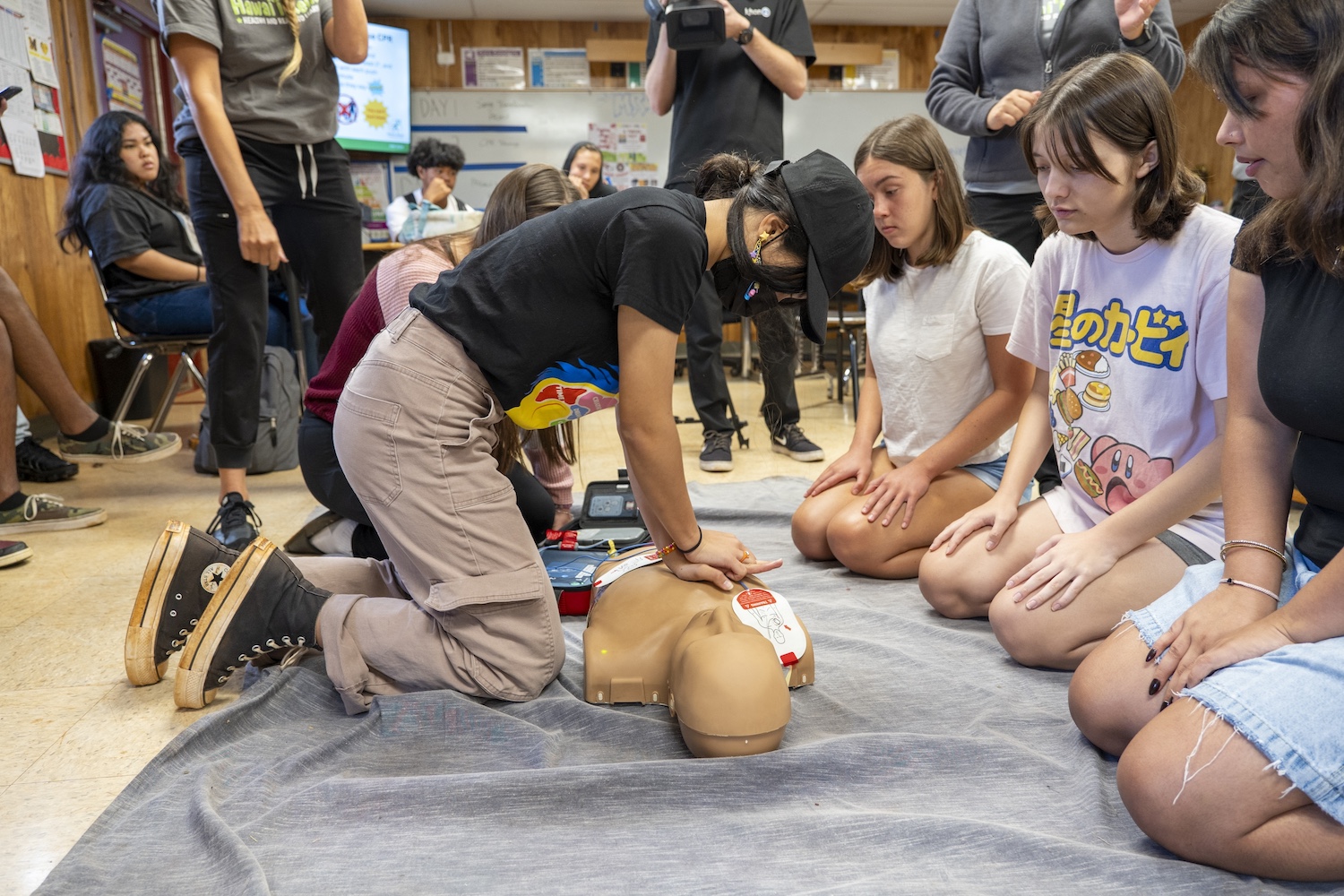 Student practicing CPR and AED on a CPR manikin. They are surrounded by other students in a classroom.