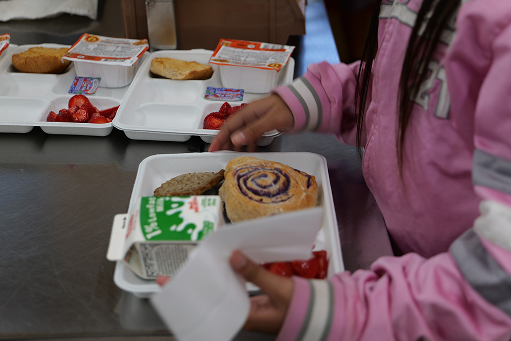 A student picks up a tray with ʻuala swirl bun, sausage patty, carton of milk and strawberries.