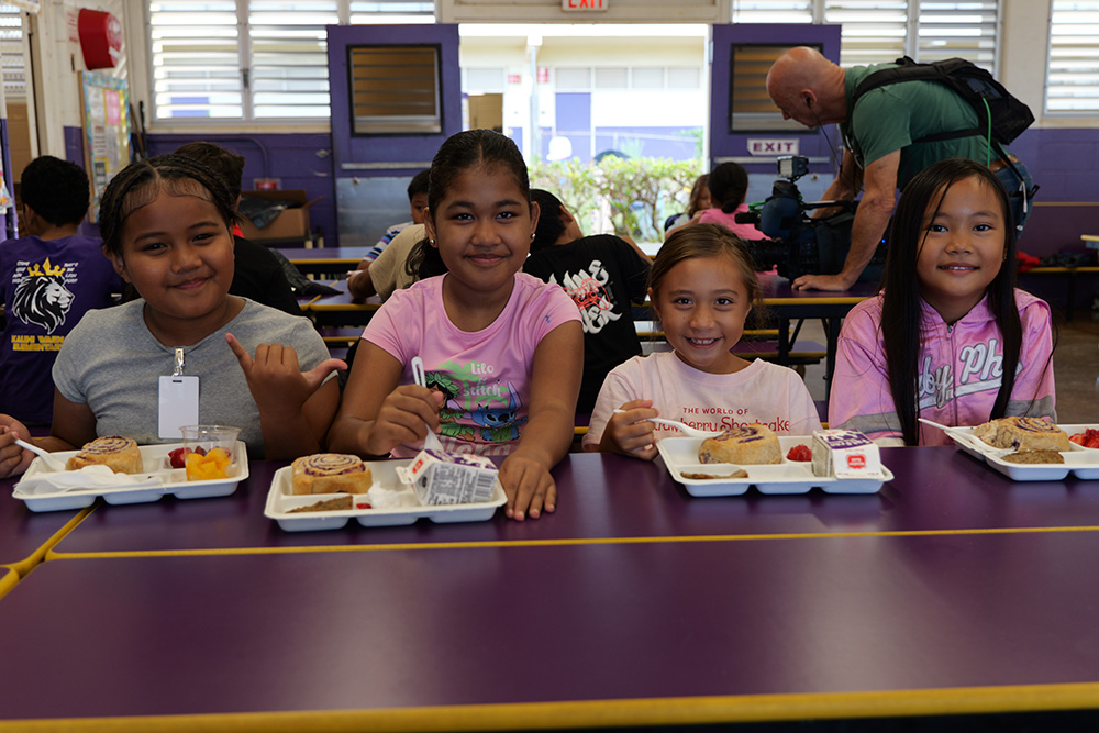 A group of students smile with their breakfast trays of ʻuala swirl bun, sausage patty, carton of milk and strawberries.