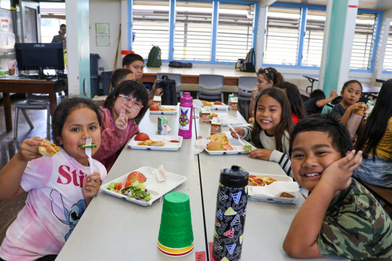 Kohala students smile with their food at a cafeteria table.