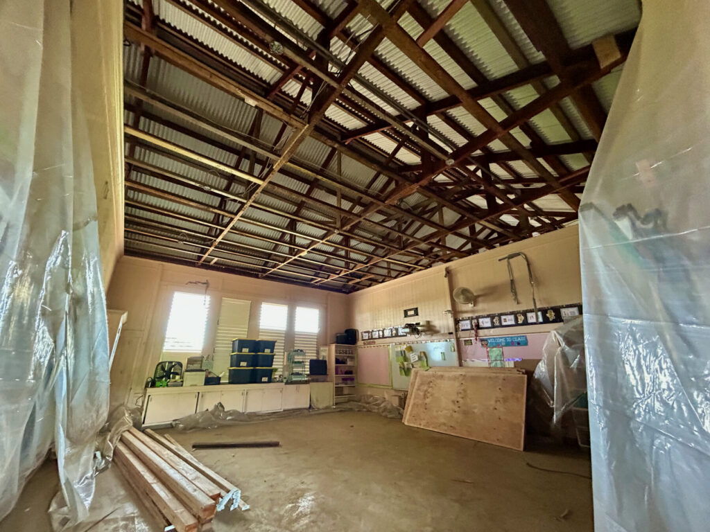 Various construction material are scattered on the ground while a ceiling has exposed trusses.
