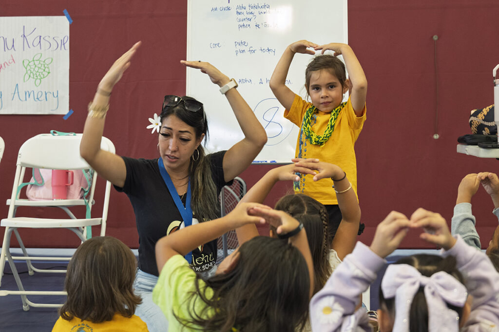 A Nā‘ālehu teacher puts her hands above her head as students follow her movement.